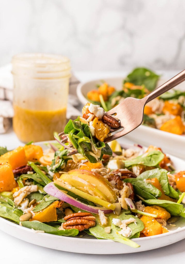 A fork lifts a bite of salad with spinach, roasted butternut squash, pecans, red onions, and dressing. A jar of dressing and a plate of salad are in the background.