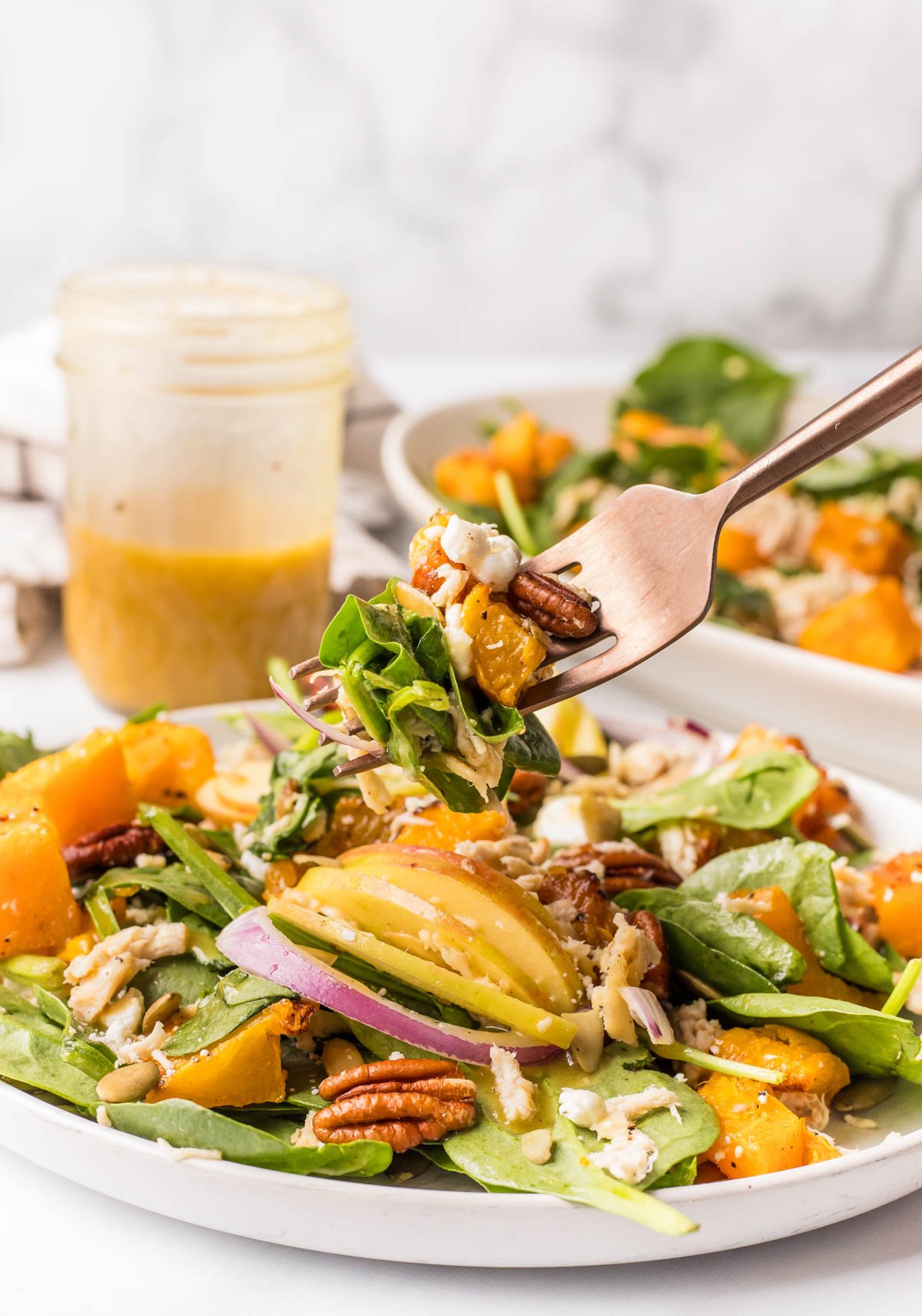A fork lifts a bite of salad with spinach, roasted butternut squash, pecans, red onions, and dressing. A jar of dressing and a plate of salad are in the background.
