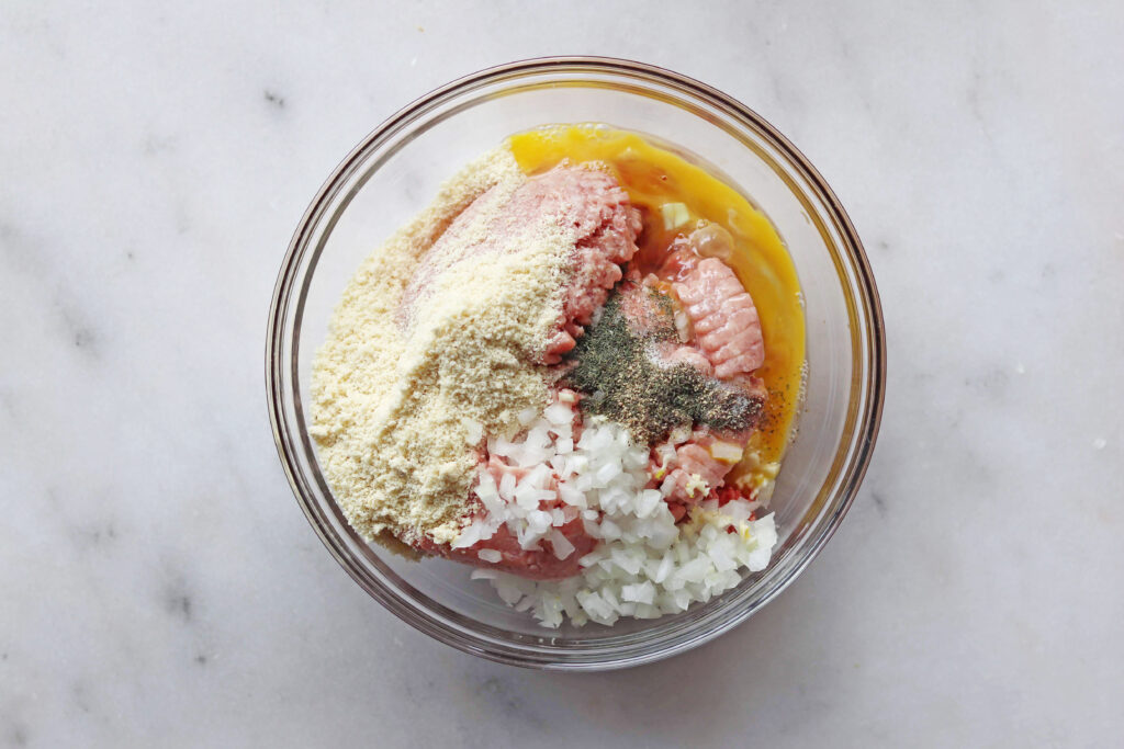 A glass bowl containing ground meat, a cracked egg, chopped onions, breadcrumbs, and seasonings—perfect for preparing Easy Swedish Meatballs on a white marble surface.