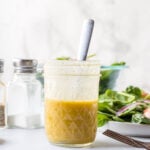 A glass jar filled with yellow salad dressing and a spoon, placed on a white surface beside a bowl of salad and salt and pepper shakers.