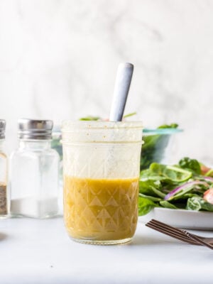 A glass jar filled with yellow salad dressing and a spoon, placed on a white surface beside a bowl of salad and salt and pepper shakers.