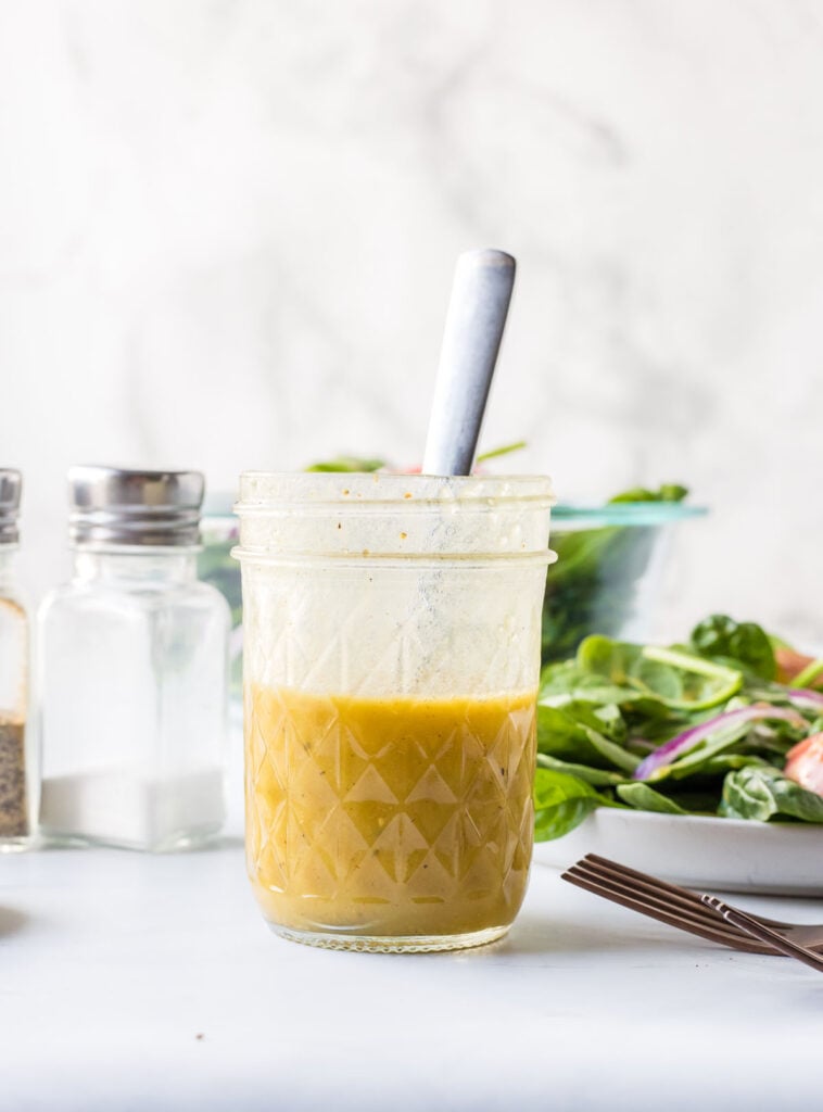 A glass jar filled with yellow salad dressing and a spoon, placed on a white surface beside a bowl of salad and salt and pepper shakers.