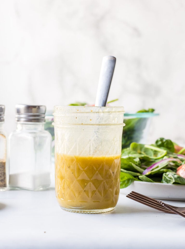 A glass jar filled with yellow salad dressing and a spoon, placed on a white surface beside a bowl of salad and salt and pepper shakers.