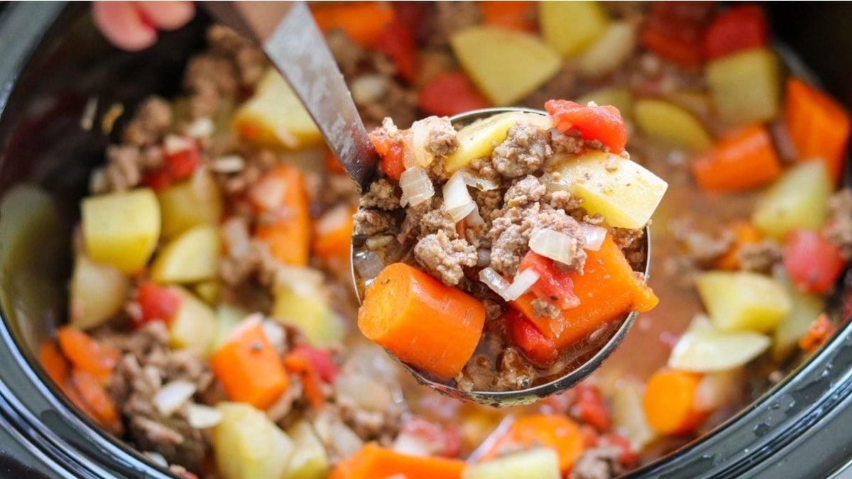A ladle lifting a serving of beef stew with carrots, potatoes, onions, and tomatoes from a pot.