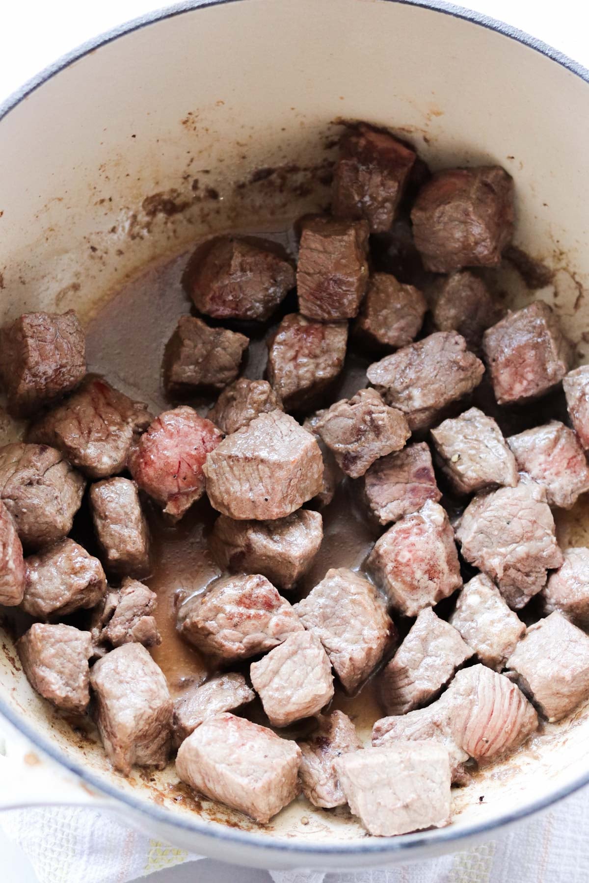 Cubed pieces of browned beef simmer in a white pot, some pieces showing a slight red center, indicating partial cooking.