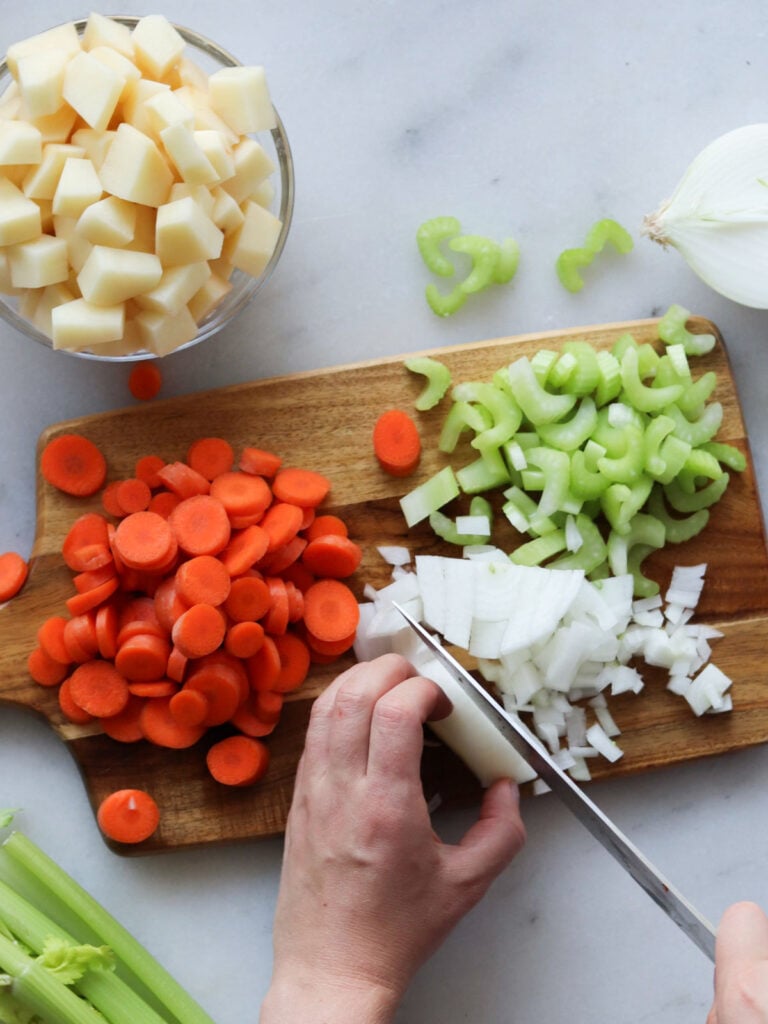 A person chopping onion on a wooden cutting board with sliced carrots, celery, and diced onion; a bowl of cubed potatoes is nearby.
