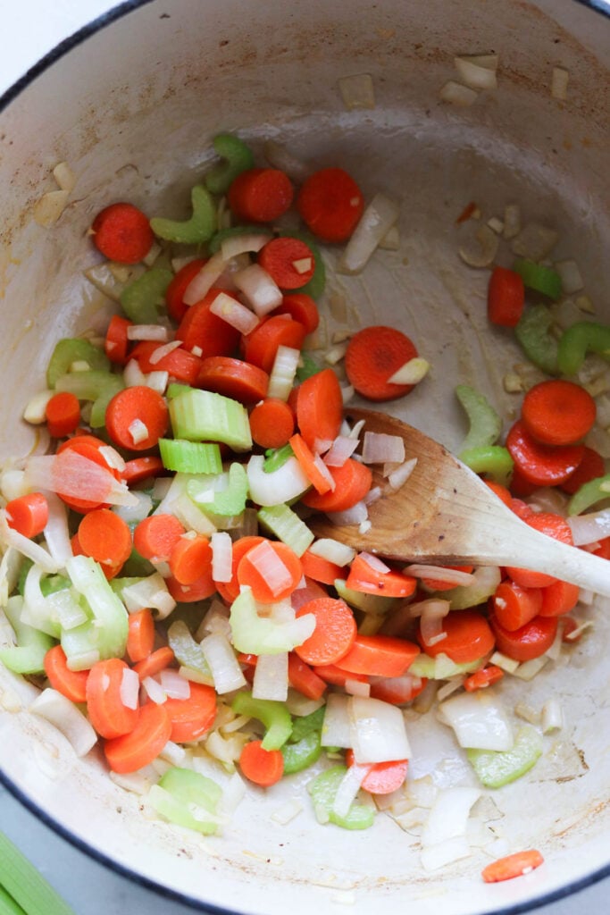 Chopped carrots, celery, and onions being sautéed in a pot with a wooden spoon.