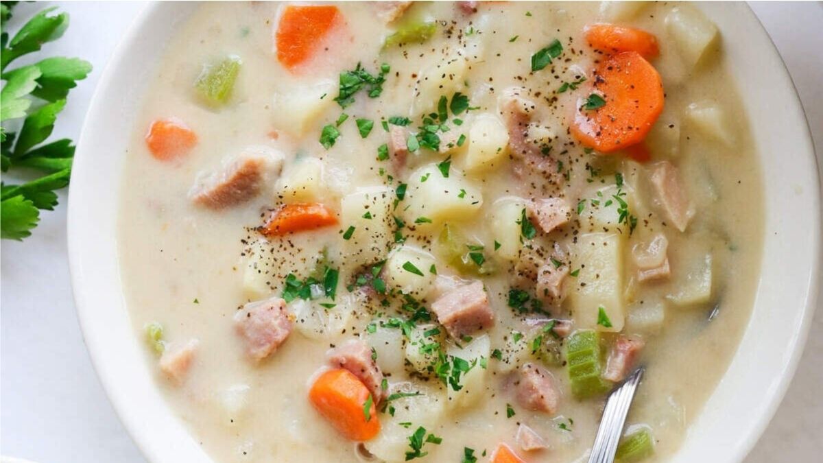 A bowl of creamy soup with chunks of ham, potatoes, carrots, and celery, garnished with chopped parsley and freshly ground black pepper. A spoon rests in the bowl.