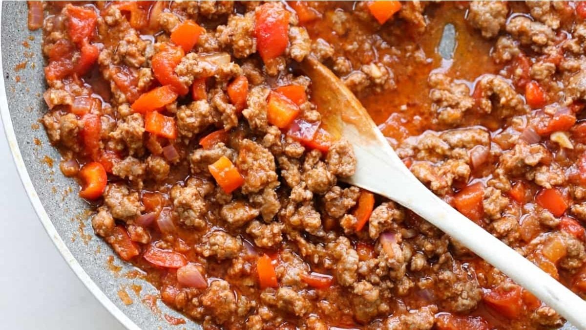 A simple dinner of ground meat with diced red bell peppers and tomato sauce in a skillet, being stirred with a wooden spoon.