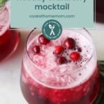 A glass of cranberry mocktail with ice, cranberries, and a sprig of rosemary makes the perfect Christmas drink, accompanied by a sign reading "holiday cranberry mocktail cookathomemom.com.