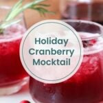 Two glasses of red cranberry mocktail with ice and rosemary garnish on a wooden surface, with text overlay: "Holiday Cranberry Mocktail"—the perfect Christmas drink for festive gatherings.