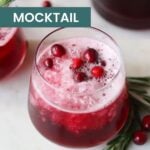 A festive cranberry mocktail, the perfect Christmas drink, is served in a glass and garnished with whole cranberries and rosemary, set on a white surface beside another glass and fresh sprigs.