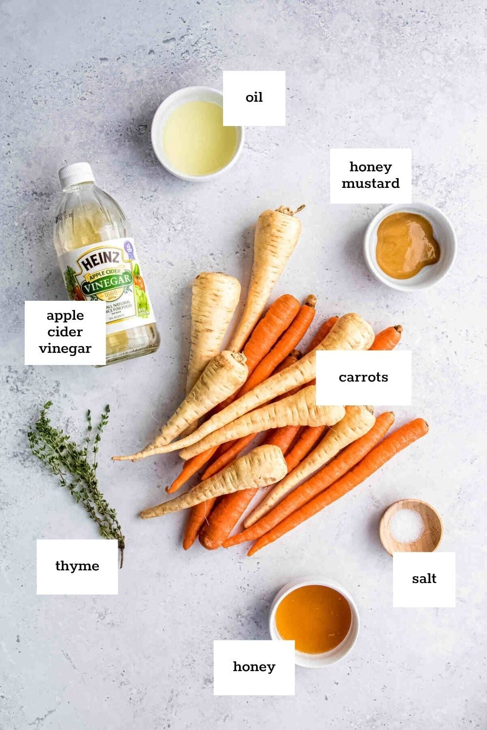 Carrots and parsnips on a surface surrounded by labeled bowls of oil, honey mustard, salt, honey, thyme, and a bottle of apple cider vinegar—perfect for making delicious honey roasted carrots.