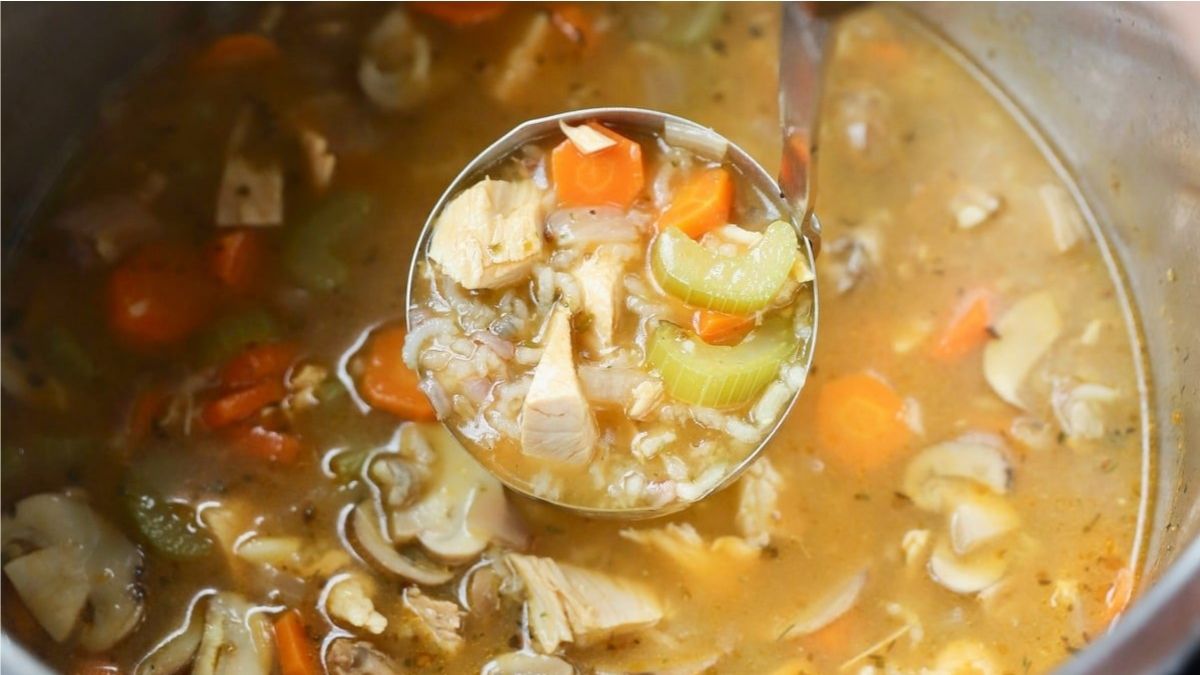 A ladle filled with chicken and vegetable soup, including pieces of chicken, carrots, celery, rice, and mushrooms, held over a pot of soup.