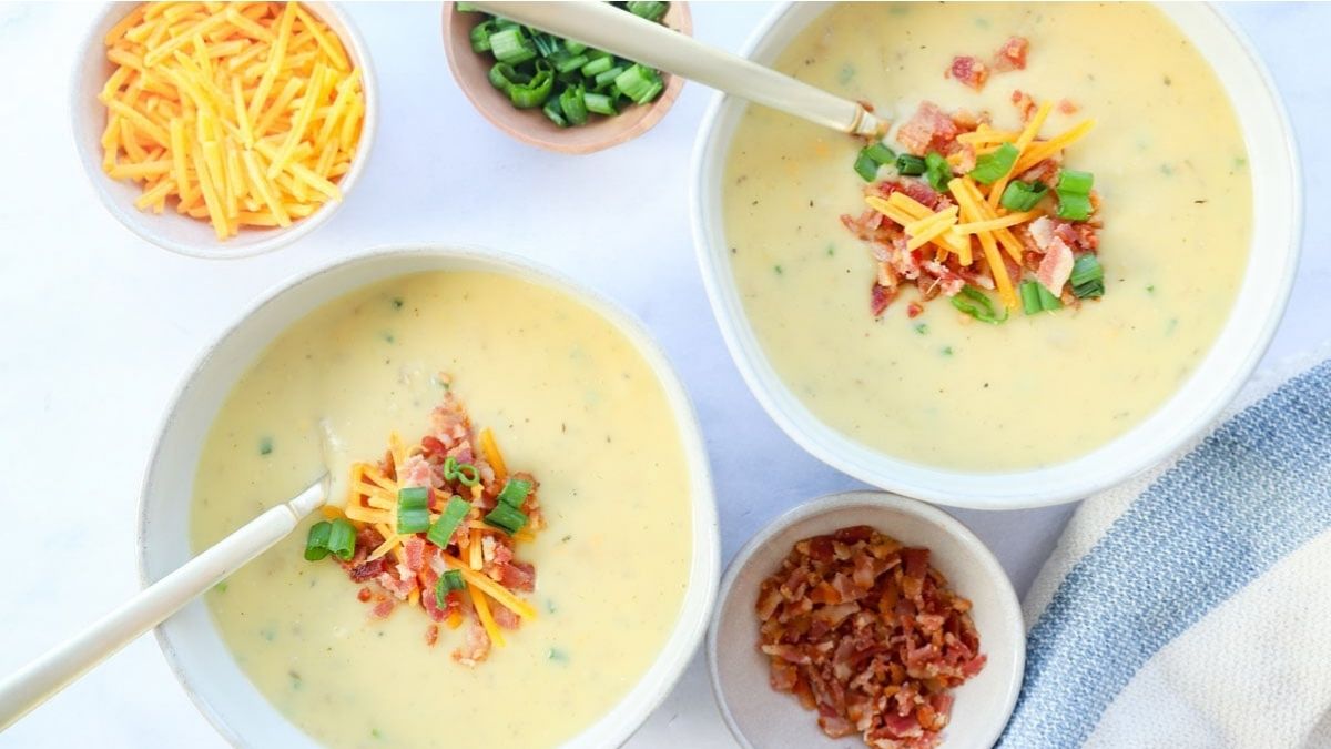Two bowls of creamy soup topped with shredded cheese, bacon bits, and chopped green onions, with small bowls of toppings on the side.