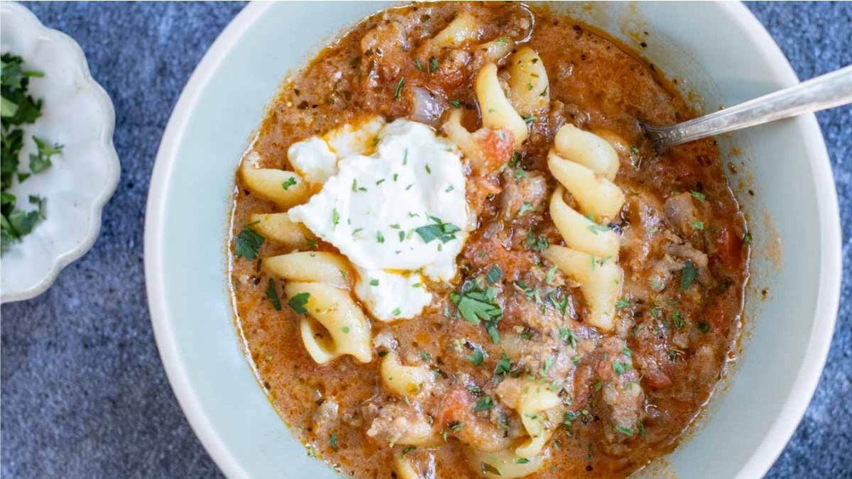 A bowl of pasta soup with spiral noodles, ground meat, tomato broth, a dollop of cream, and chopped herbs, with a spoon in the bowl.