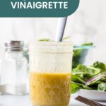 A glass jar of maple vinaigrette with a spoon sits on the counter, surrounded by fresh greens and a salt shaker. The label reads "Maple Vinaigrette," highlighting this flavorful dressing.