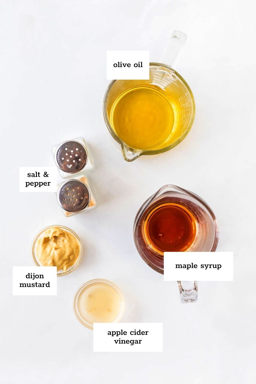 Overhead view of labeled ingredients for a maple vinaigrette: olive oil, maple syrup, dijon mustard, apple cider vinegar, salt, and pepper.