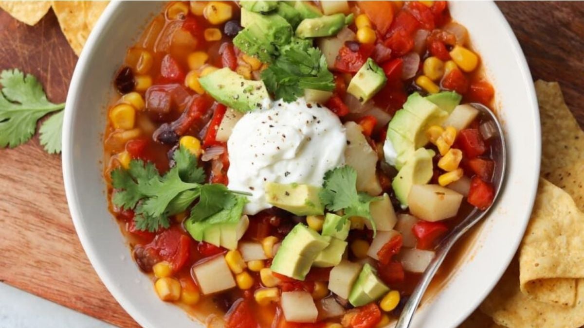 A bowl of vegetable soup topped with diced avocado, cilantro, and a dollop of sour cream, with a spoon and tortilla chips on the side.