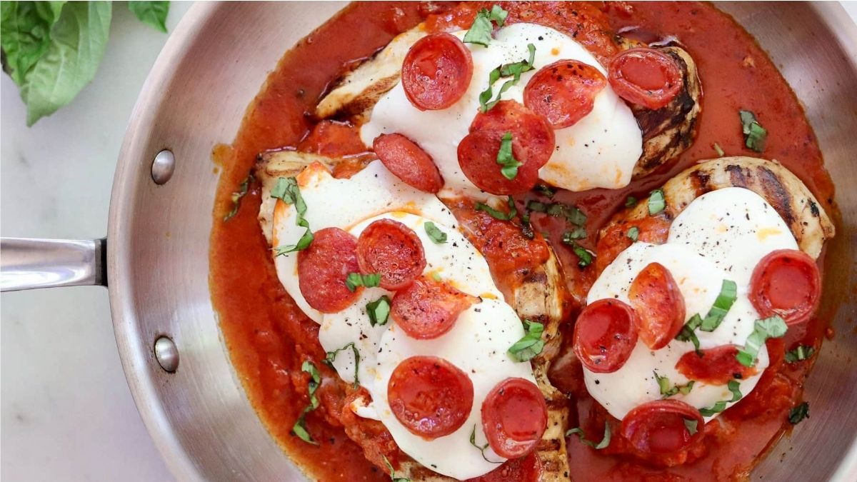 Three grilled chicken breasts in tomato sauce topped with melted mozzarella, pepperoni slices, and chopped fresh basil in a metal pan.