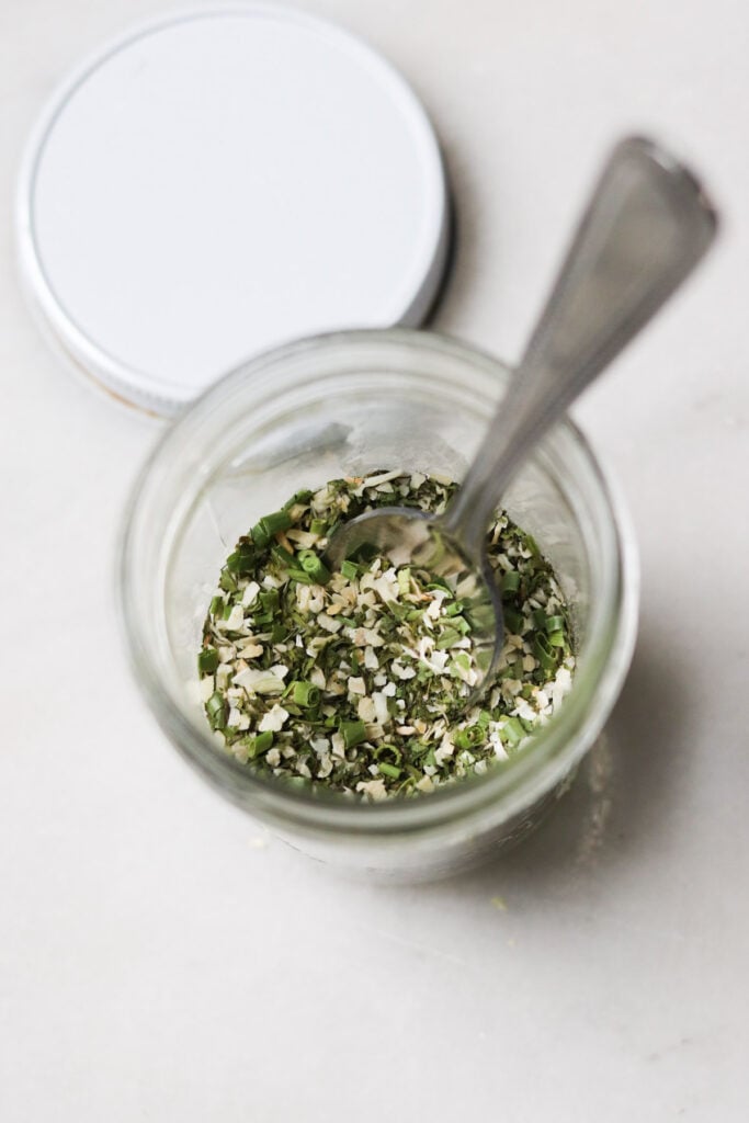 A glass jar filled with dried herbs and seasonings, with a spoon inside and the lid resting nearby on a light surface.
