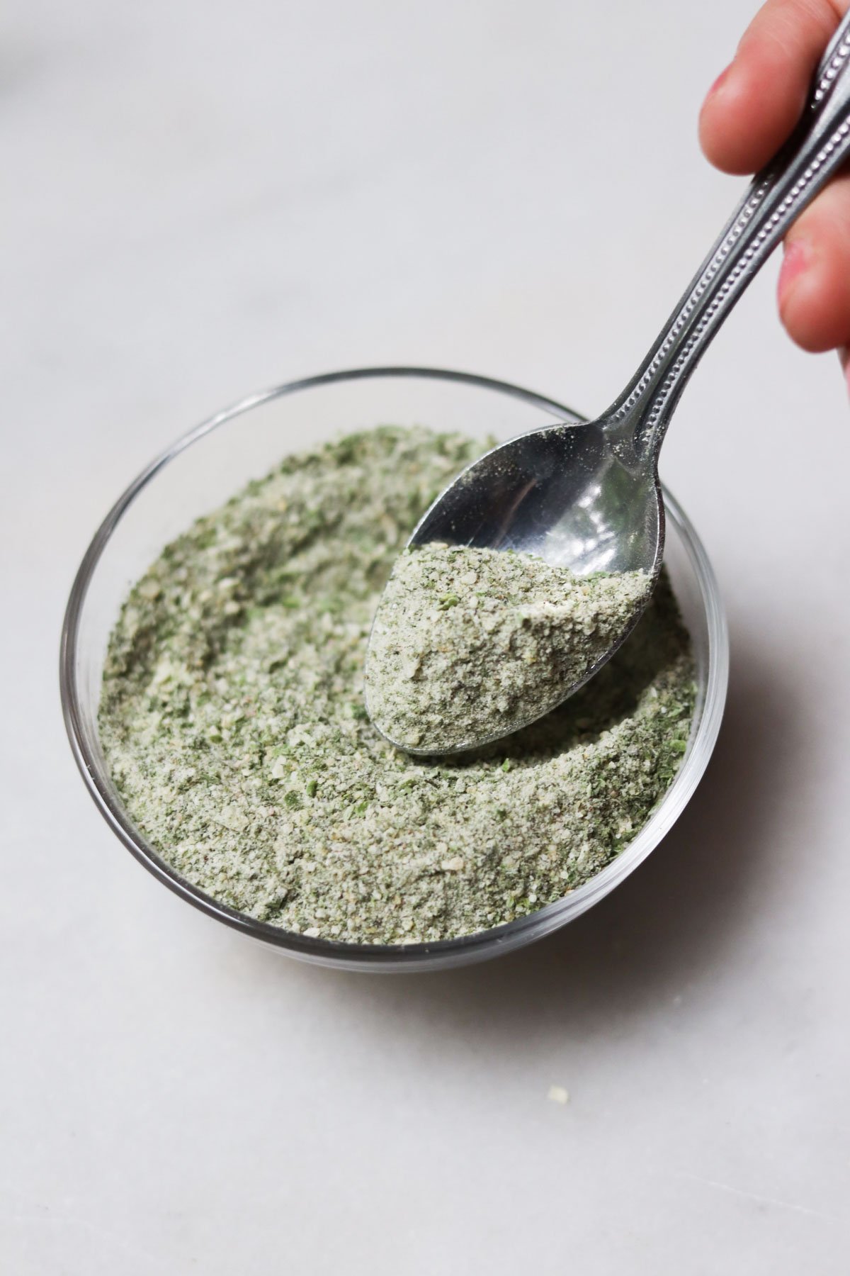 A hand holds a spoon scooping a powdered seasoning mix from a small clear glass bowl on a white surface.