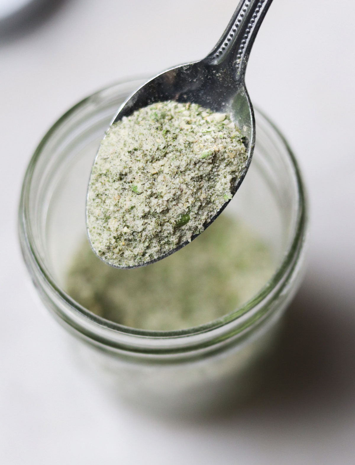 A metal spoon holding a green and white powder mixture above an open glass jar filled with the same powder.