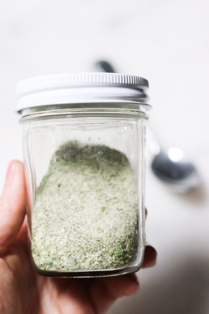 A hand holding a glass jar filled with a greenish dried seasoning mix; a spoon and white surface are blurred in the background.