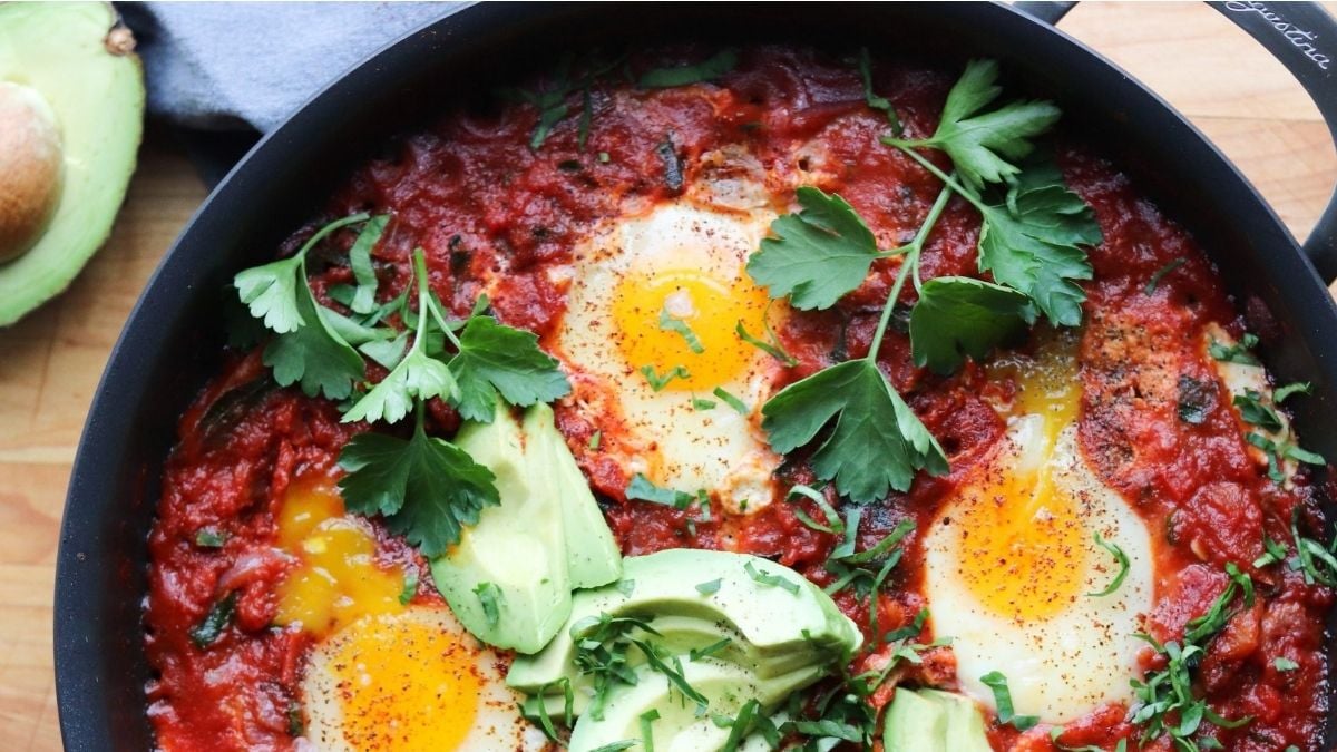 A skillet with three eggs cooked in a tomato sauce, garnished with fresh parsley and sliced avocado on a wooden surface.