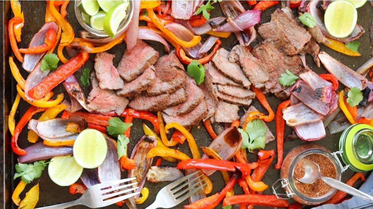 Sliced grilled steak with red and yellow bell peppers, red onions, lime wedges, and fresh cilantro on a tray, with seasoning and two forks nearby.