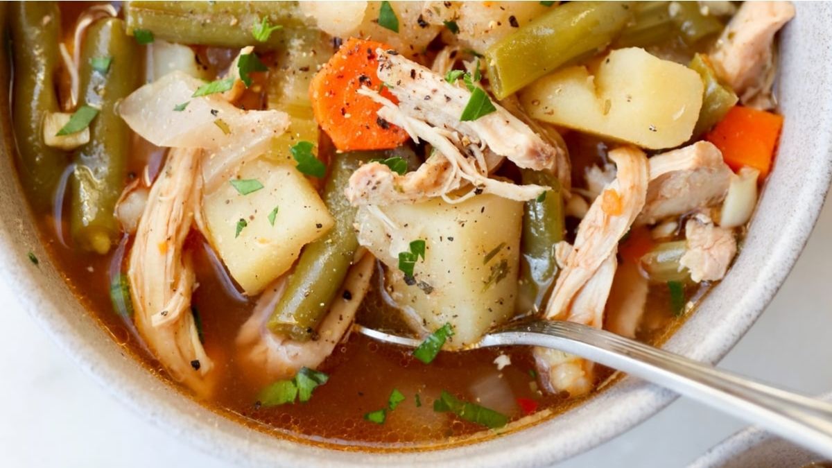 A bowl of chicken stew with shredded chicken, potatoes, green beans, carrots, and herbs, with a spoon inside the bowl.