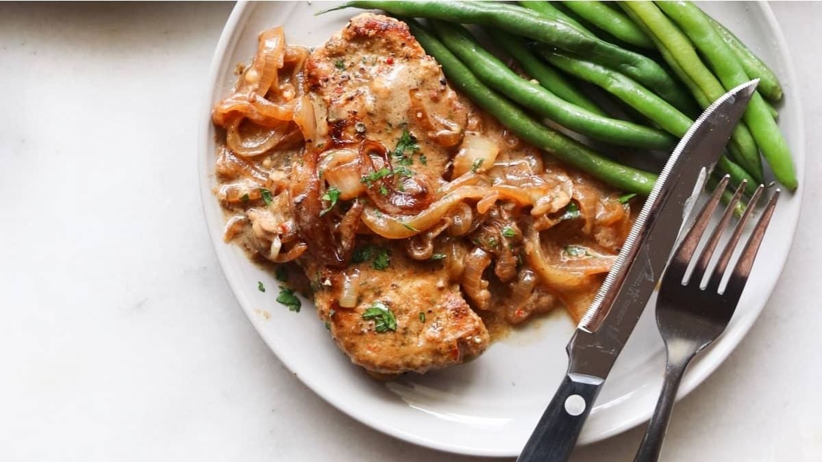 A plate with smothered pork chops topped with sautéed onions and sauce, served with green beans. A fork and knife are placed beside the plate.