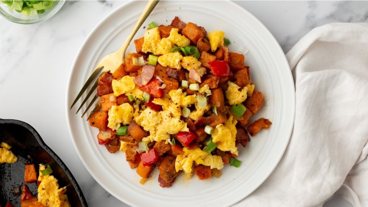 A white plate with scrambled eggs, diced sweet potatoes, red bell peppers, onions, and green onions, with a gold fork on the side.