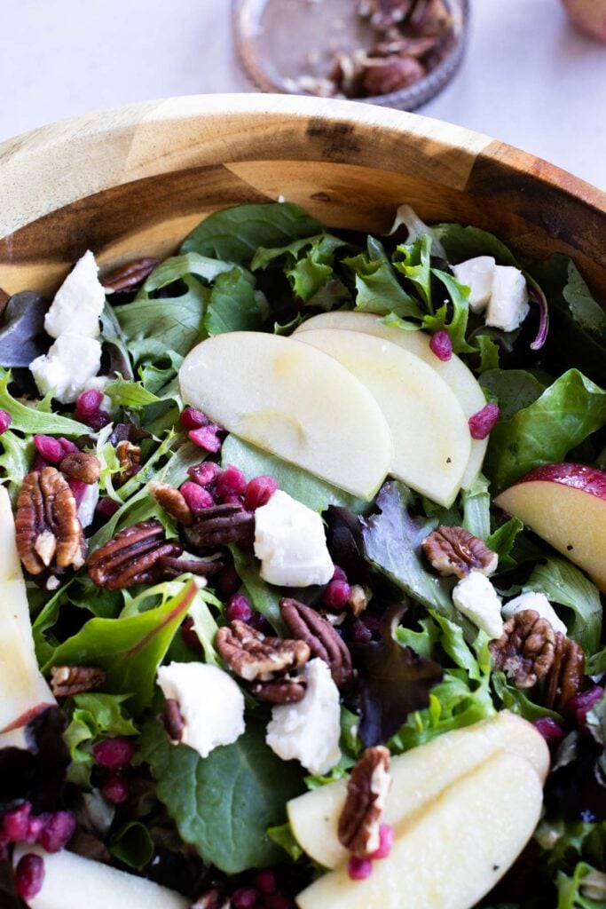 A salad with mixed greens, sliced apples, pecans, goat cheese, and pomegranate seeds in a wooden bowl.