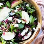 A wooden bowl filled with mixed greens, apple slices, pecans, pomegranate seeds, and crumbled cheese, with a light brown cloth nearby.