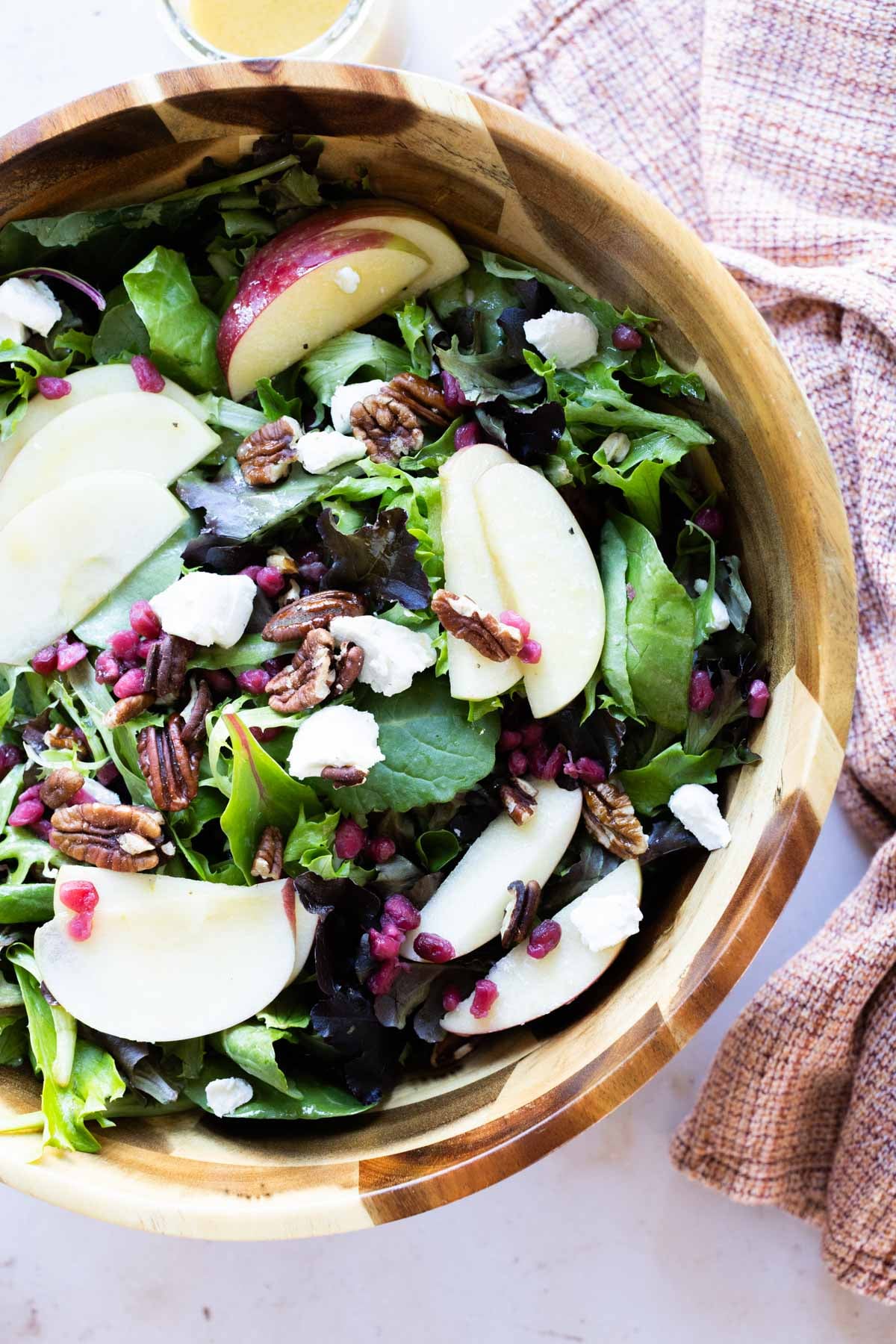 A wooden bowl filled with a holiday salad of mixed greens, apple slices, pecans, pomegranate seeds, and crumbled cheese, with a light brown cloth napkin nearby.