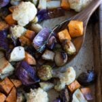 A wooden spoon scoops roasted vegetables, including red onion, cauliflower, Brussels sprouts, sweet potato, and herbs, from a baking sheet.