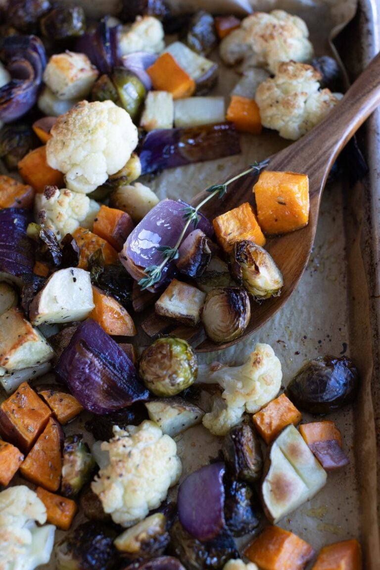A wooden spoon scoops roasted vegetables, including red onion, cauliflower, Brussels sprouts, sweet potato, and herbs, from a baking sheet.