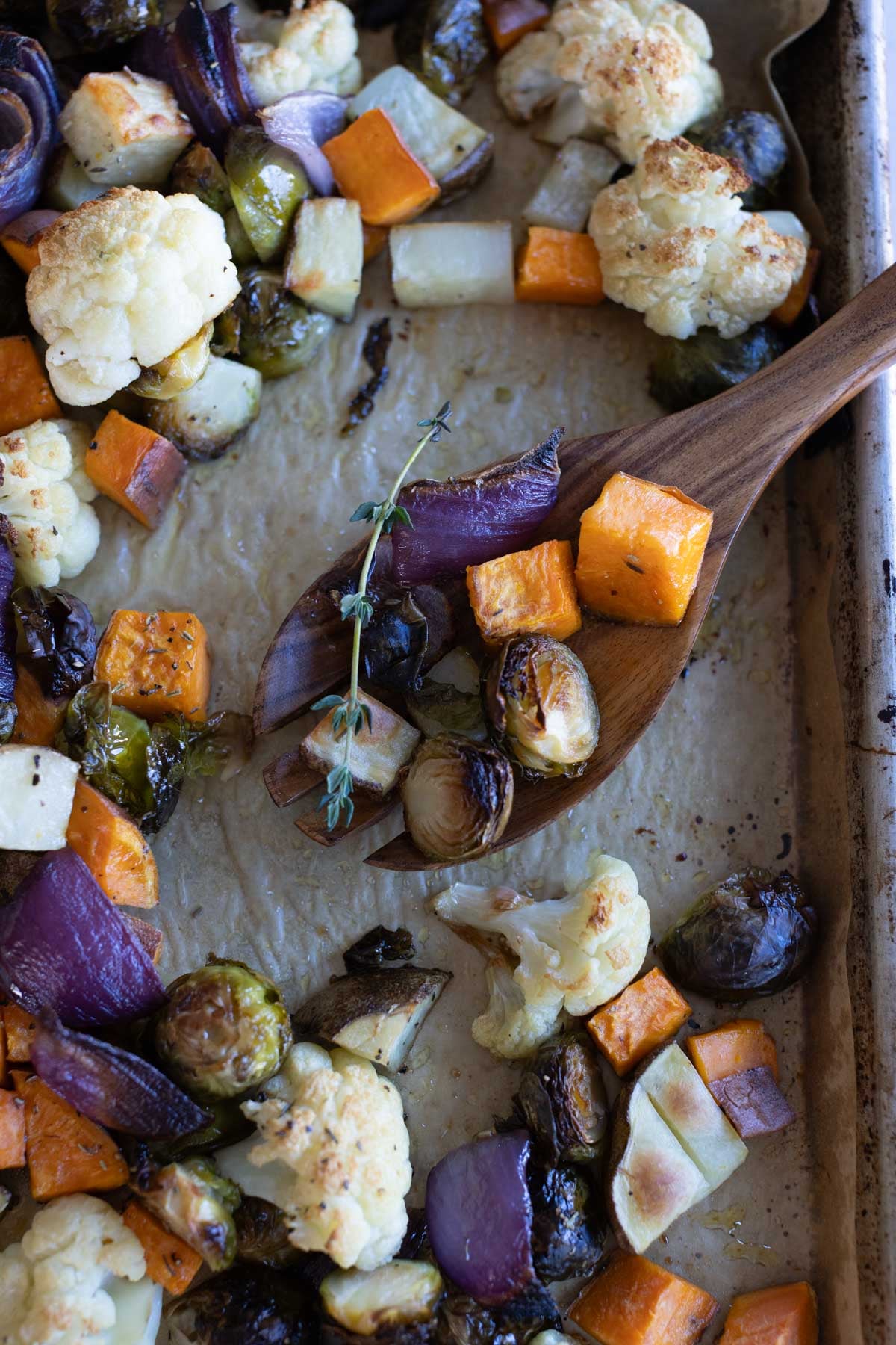 A baking sheet with roasted vegetables including cauliflower, Brussels sprouts, red onion, and sweet potato, with a wooden spoon and a sprig of thyme.