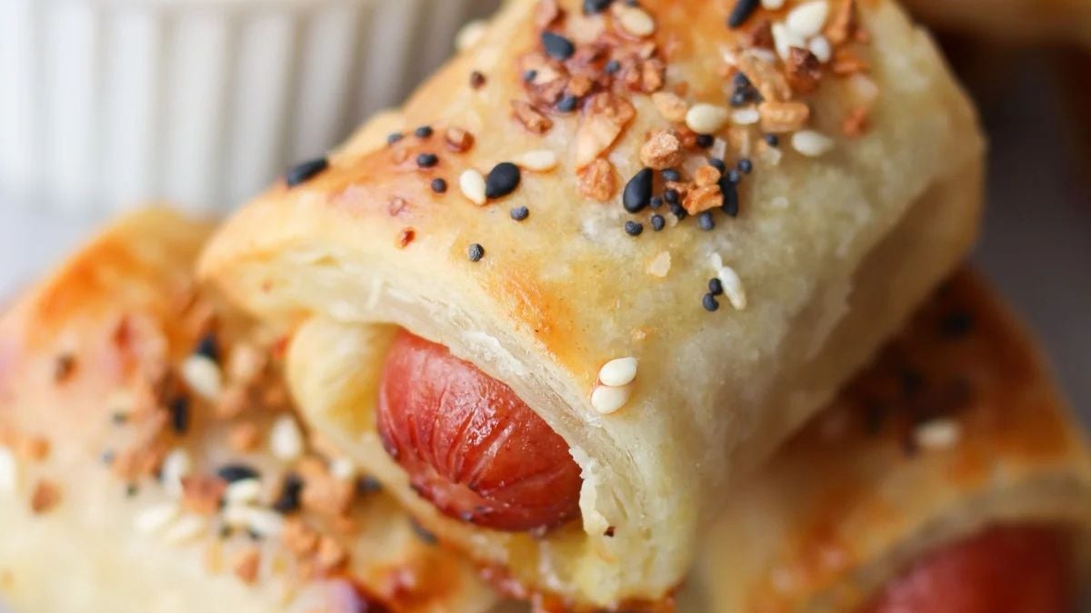 Close-up of a pastry-wrapped sausage topped with sesame seeds and seasonings, with a white container visible in the background.