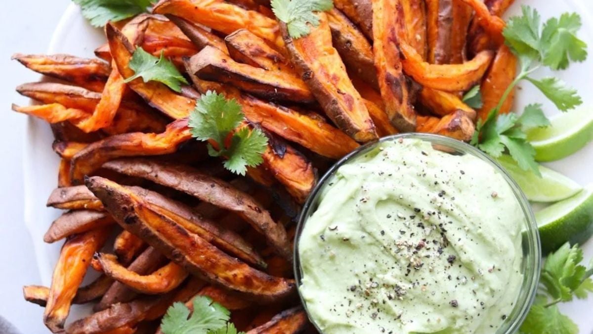 A plate of baked sweet potato fries garnished with cilantro, served with a bowl of creamy green dip and lime wedges.
