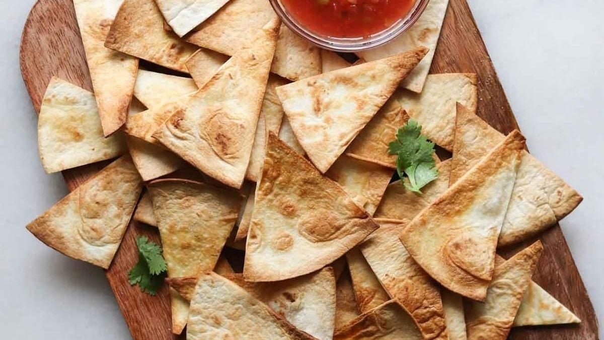 Baked tortilla chips arranged on a wooden board with a small bowl of red salsa and a few cilantro leaves.