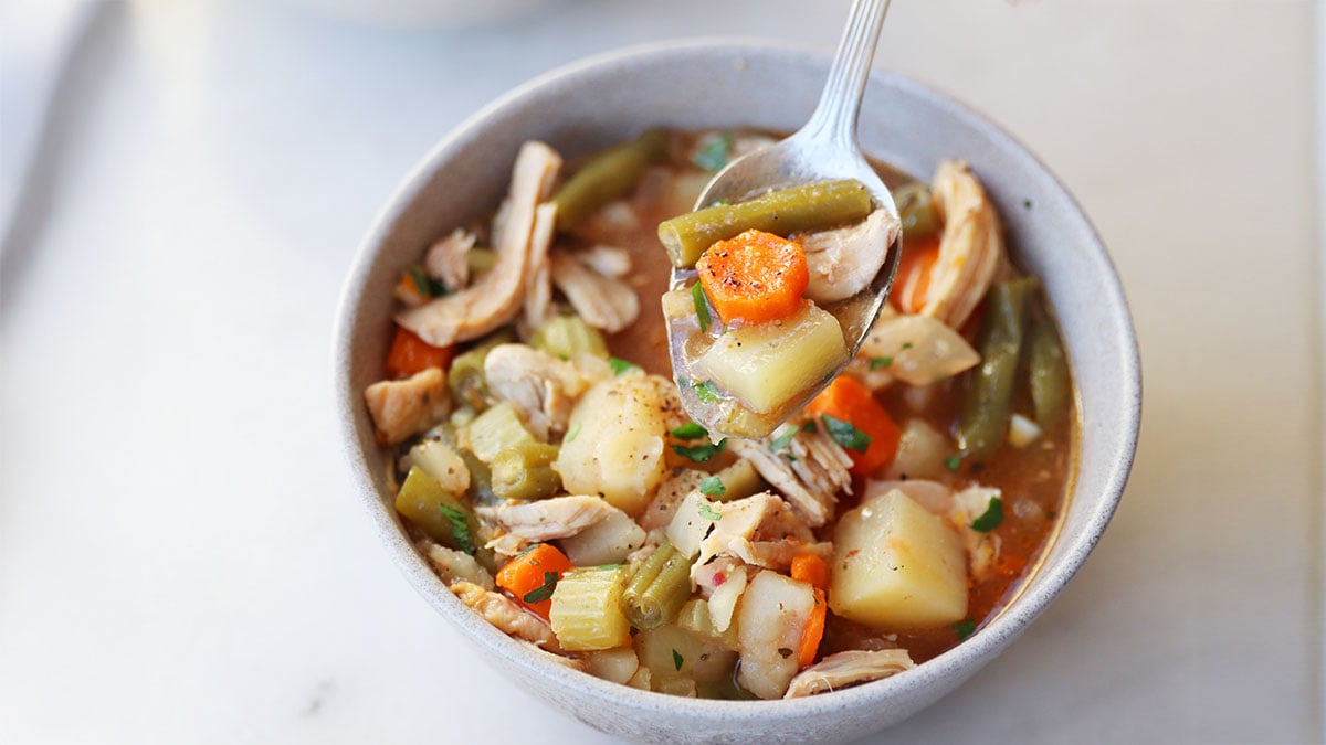 A bowl of chicken and vegetable stew with potatoes, carrots, green beans, and celery; a spoon lifts a portion from the bowl.