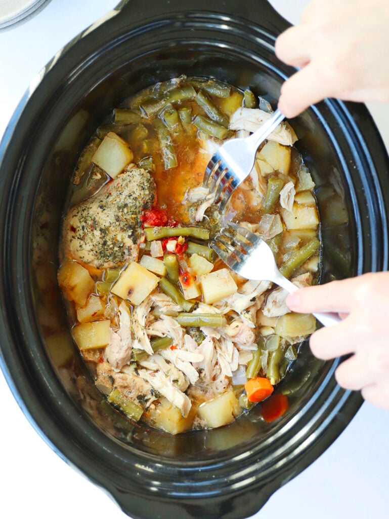 A person uses two forks to shred cooked chicken in a slow cooker with green beans, potatoes, and carrots in broth.