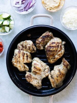 Grilled chicken pieces in a round grill pan, surrounded by bowls of sliced red onion, hummus, feta cheese, cucumber, and tomato.