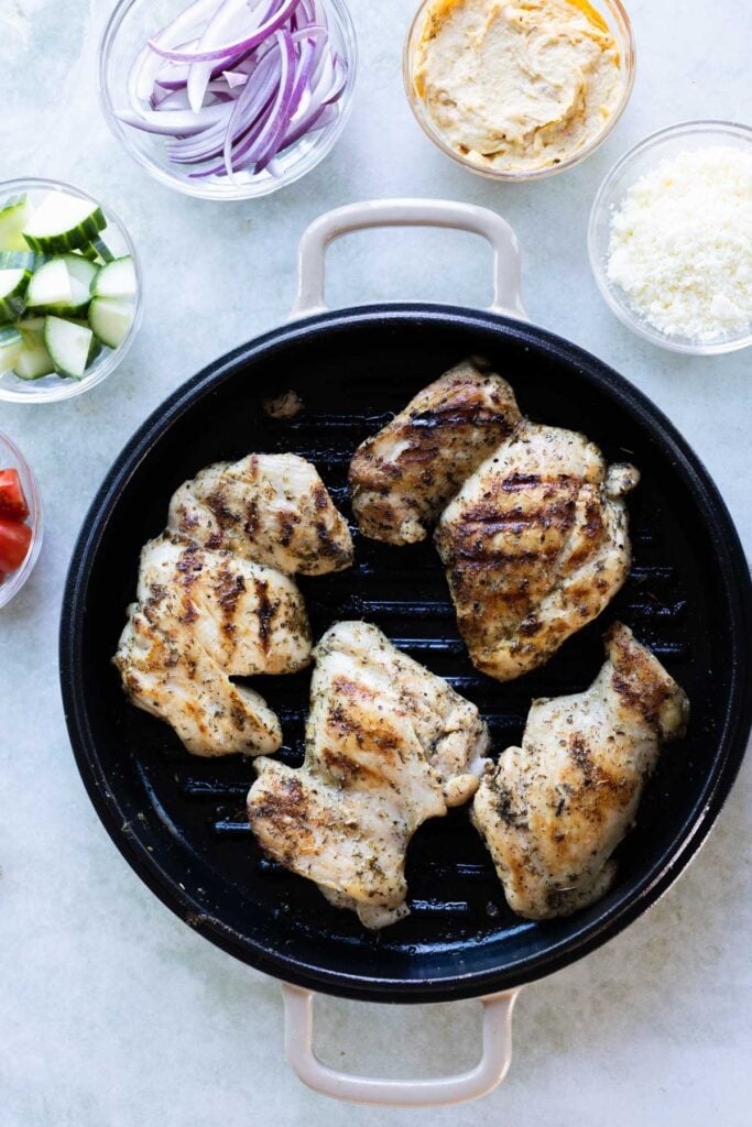Grilled chicken pieces in a round grill pan, surrounded by bowls of sliced red onion, hummus, feta cheese, cucumber, and tomato.