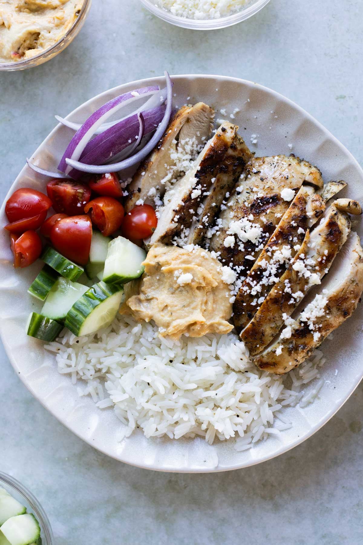 A plate with sliced grilled chicken, white rice, cucumber, tomatoes, red onion, hummus, and crumbled feta cheese.