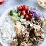 A plate with grilled chicken pieces, white rice, cucumber, cherry tomatoes, red onion, hummus, and feta cheese. Bowls of onion, tomato, and hummus are in the background.