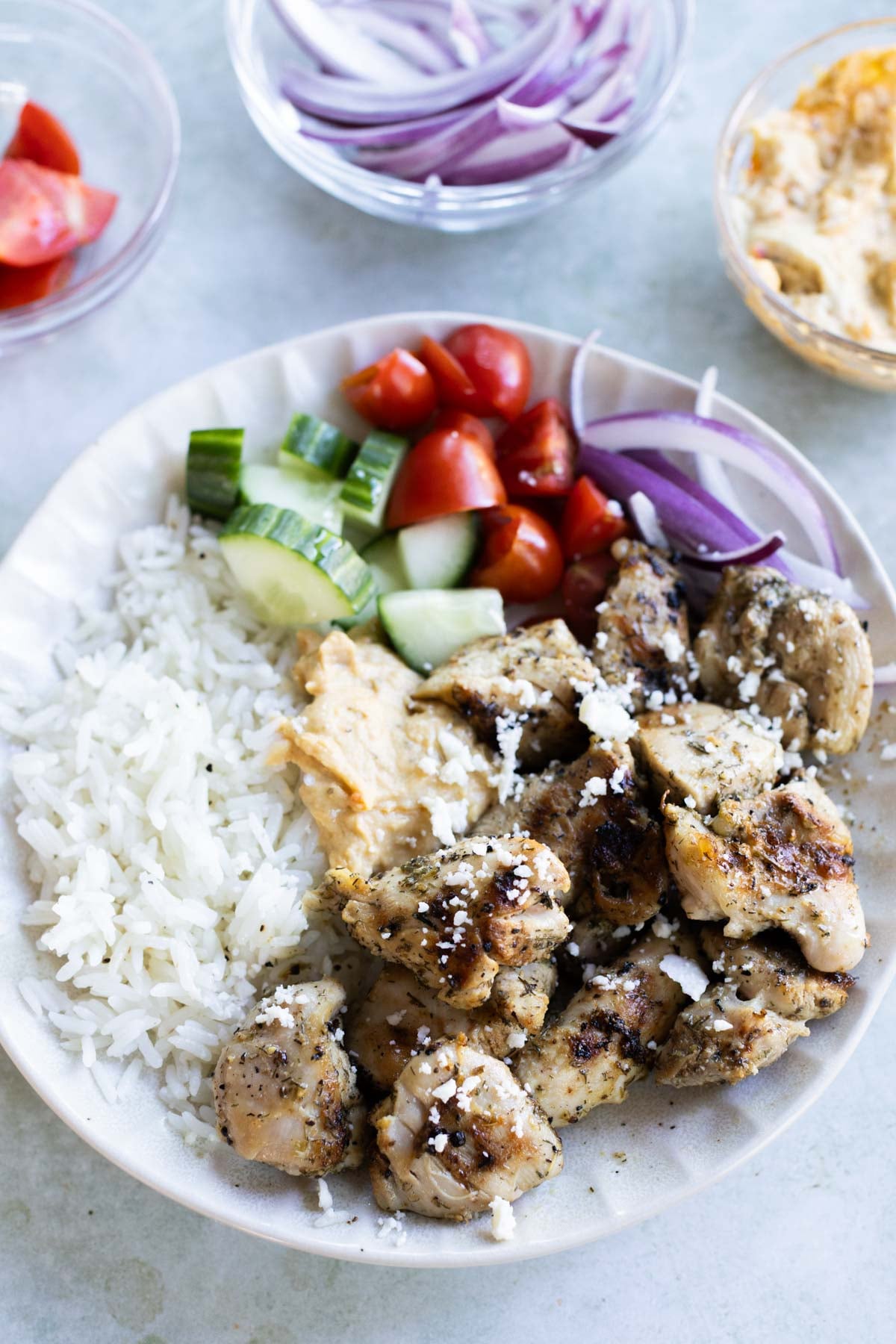 A plate with grilled chicken pieces, white rice, cucumber, cherry tomatoes, red onion, hummus, and feta cheese. Bowls of onion, tomato, and hummus are in the background.