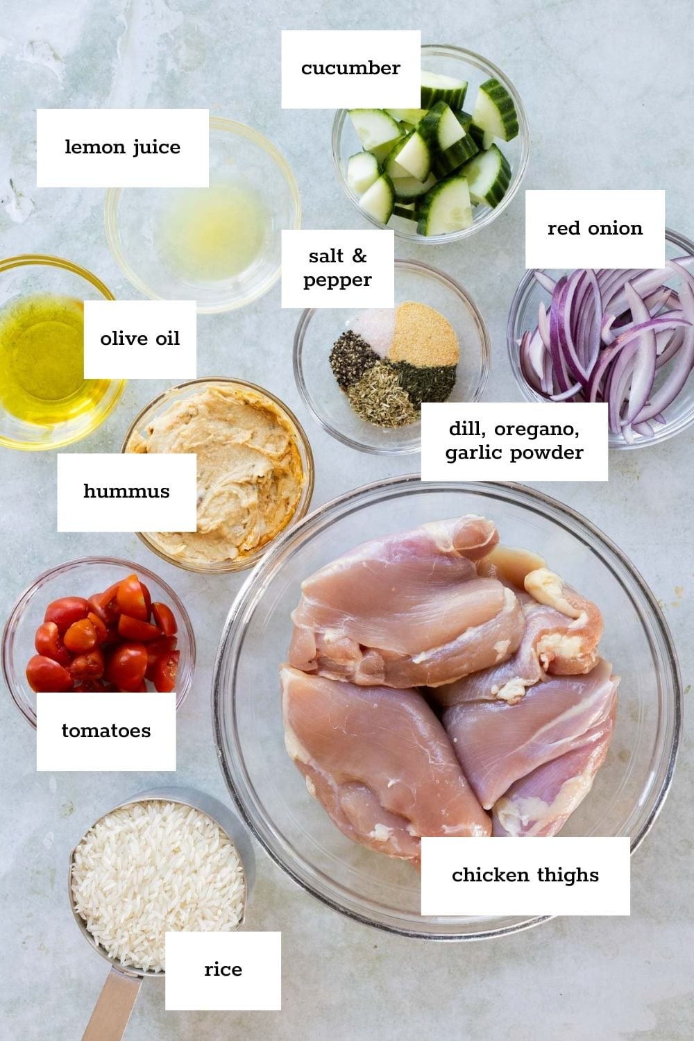Top-down view of labeled ingredients for a chicken dish, including chicken thighs, rice, tomatoes, hummus, cucumber, red onion, lemon juice, olive oil, seasonings, salt, and pepper.